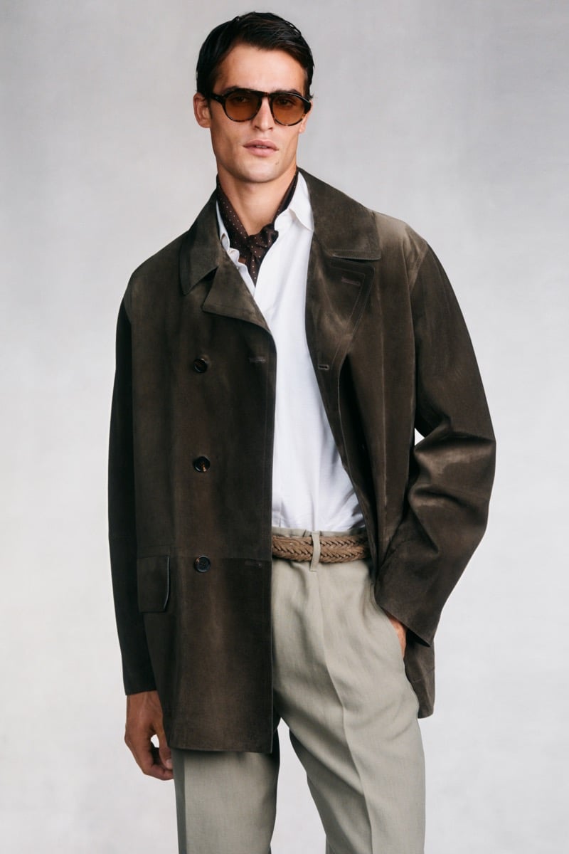 Model Parker Van Noord in tortoise aviator sunglasses wears a dark brown suede car coat over a white shirt and brown printed neckerchief, paired with sage green pleated trousers and a woven tan belt