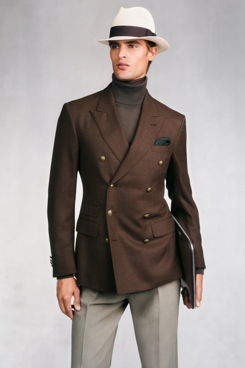 Model Parker Van Noord in a cream Panama hat with a brown band wears a brown double-breasted blazer with gold buttons and dark green pocket square over a brown turtleneck and printed scarf, paired with gray trousers and a brown leather portfolio