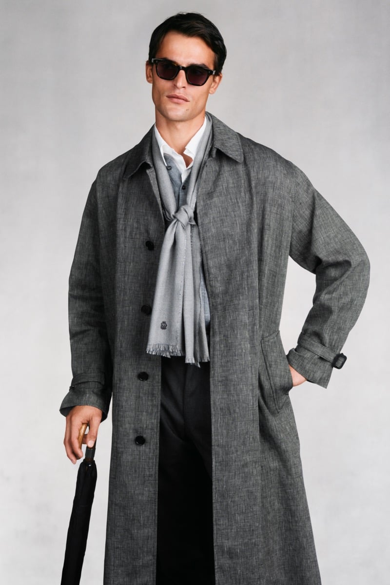 Model Parker Van Noord in dark sunglasses wears a gray linen single-breasted trench coat over a gray suit, white shirt, and gray fringed scarf, holding a black umbrella