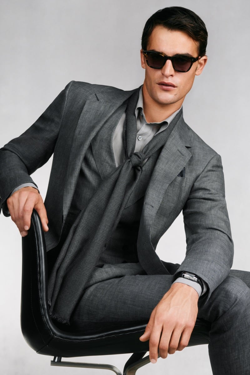 Model Parker Van Noord in tortoise sunglasses sits in a black leather chair in a charcoal high-twist linen three-piece suit with peak lapels over a gray polo and gray knotted scarf