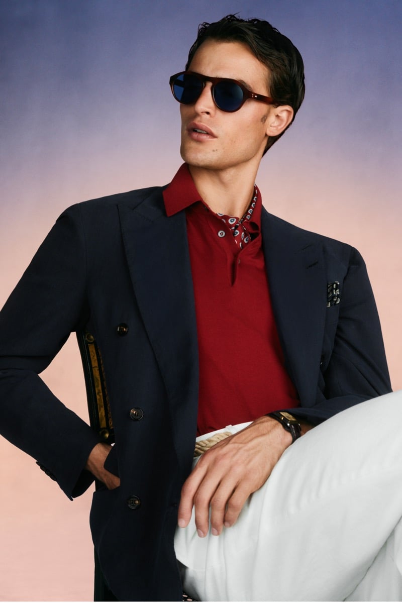 Model Parker Van Noord in tortoise round sunglasses wears a navy unstructured double-breasted blazer over a red polo and printed silk scarf, paired with white trousers and a rope belt