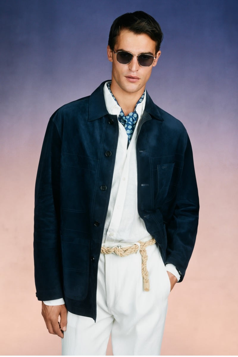 Model Parker Van Noord in slim metal sunglasses wears a navy suede chore jacket over a white shirt and blue printed neckerchief, paired with white trousers and a sculpted rope belt