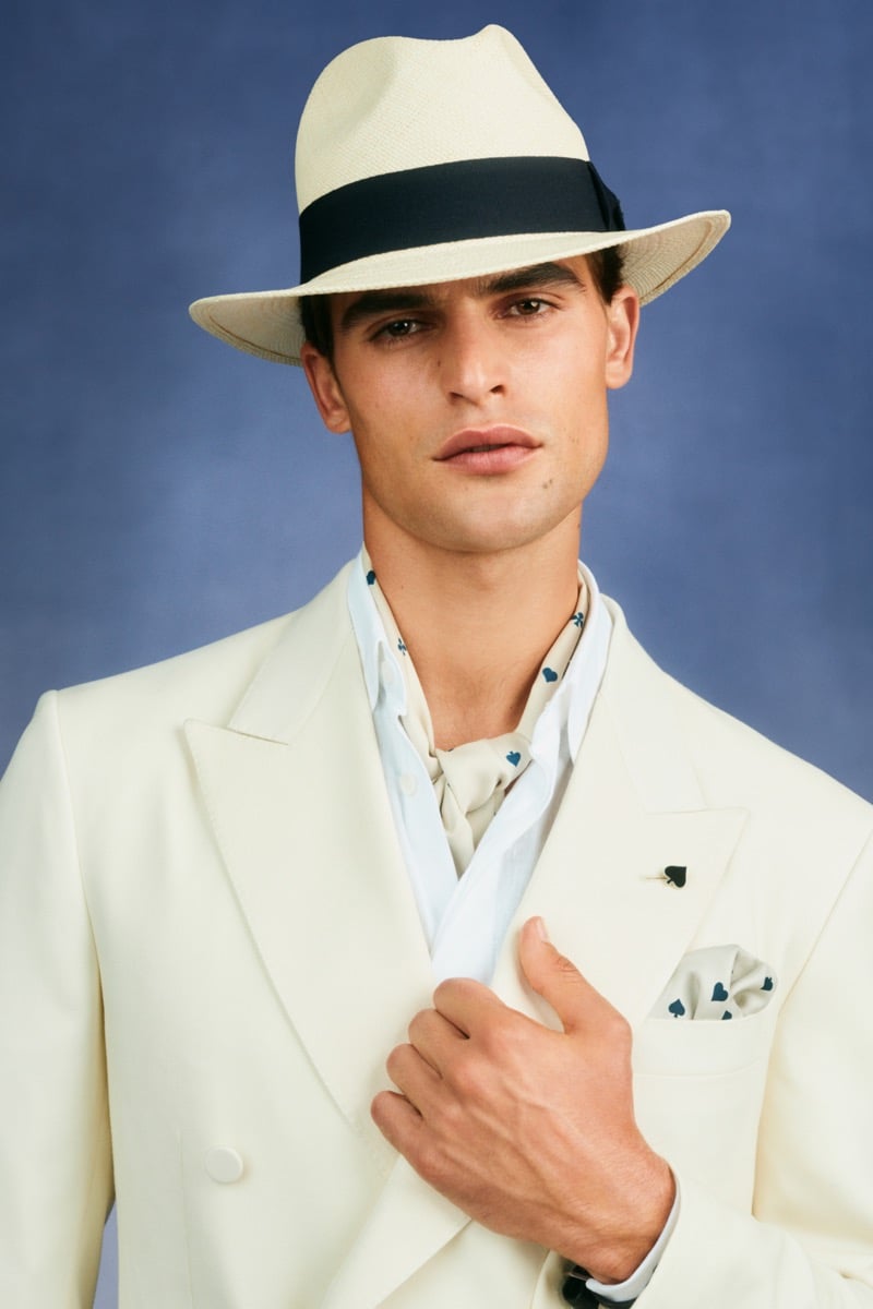 Model Parker Van Noord in a cream Panama hat with a navy band wears a cream double-breasted blazer with a playing-card spade lapel pin and printed silk pocket square over a pale blue club-print shirt and cream silk scarf