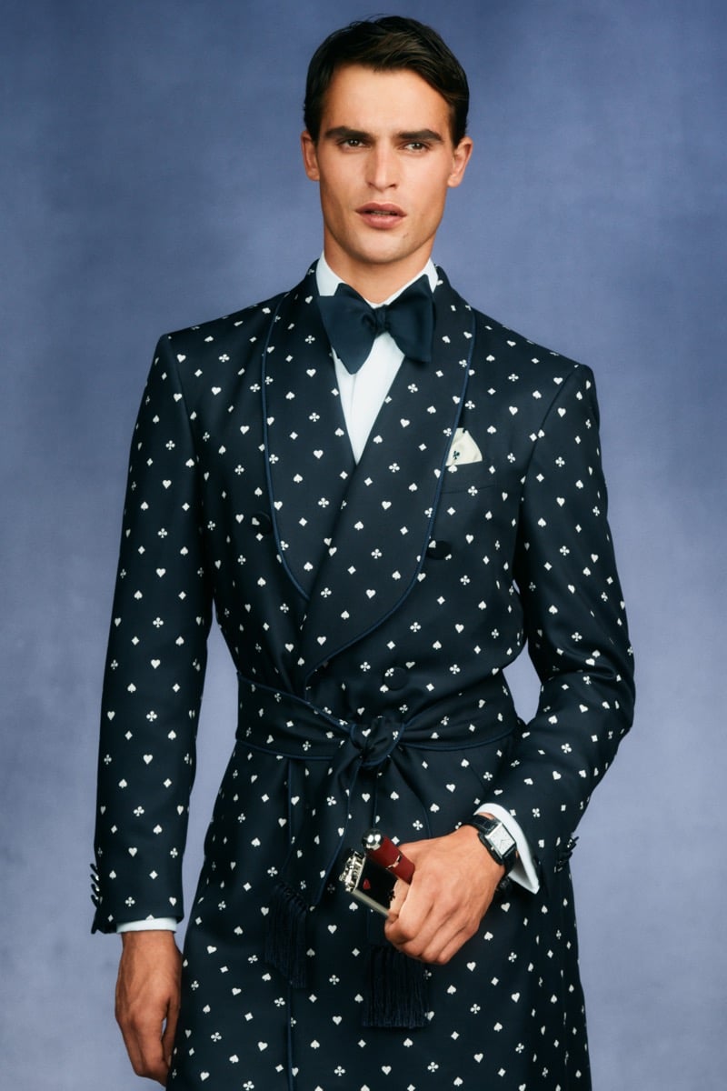 Model Parker Van Noord in a navy silk jacquard tuxedo robe printed with white playing-card motifs, layered over a white shirt and navy bow tie, with an Ace of Hearts playing-card lighter in hand