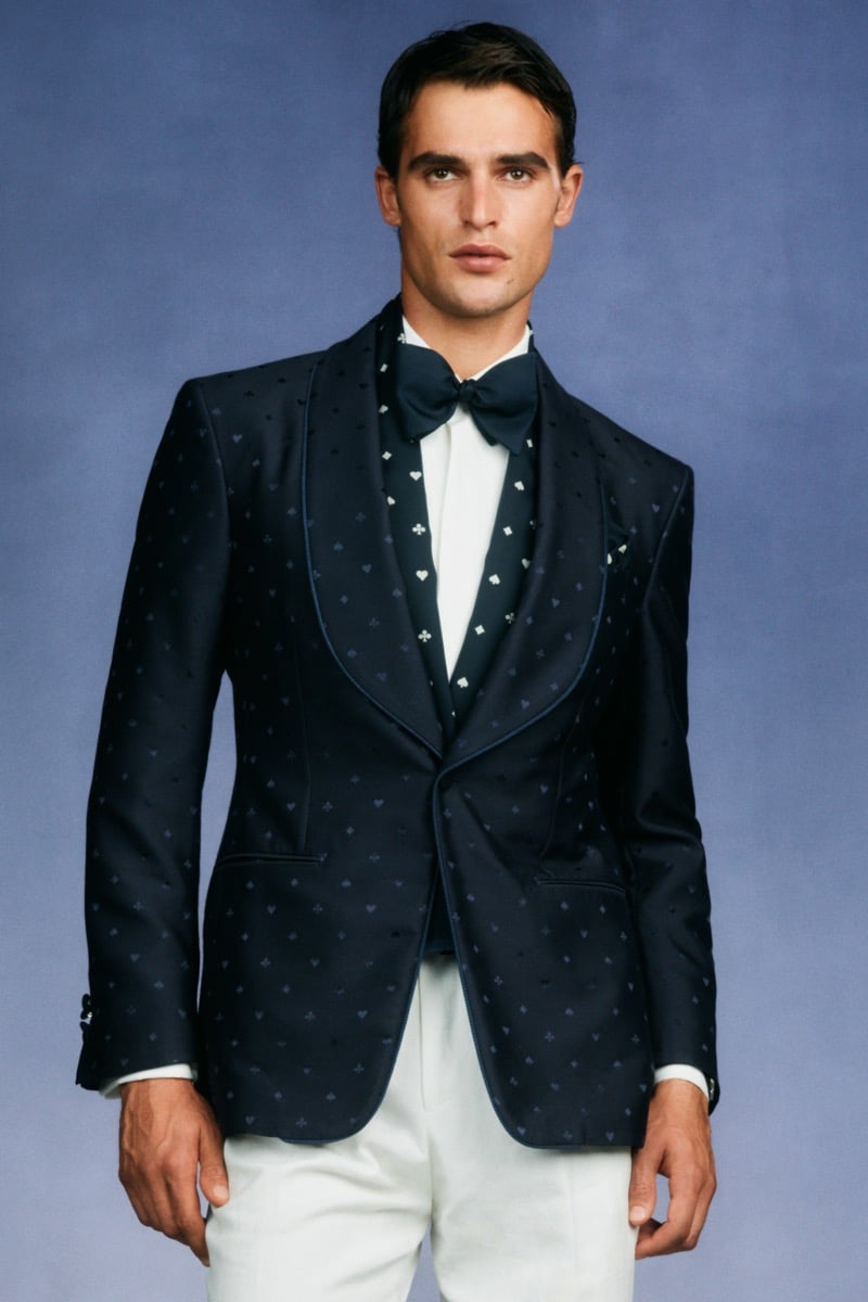 Model Parker Van Noord in a navy shawl-collar dinner jacket with tonal playing-card jacquard over a navy waistcoat printed with playing-card motifs, a white shirt, and a navy bow tie