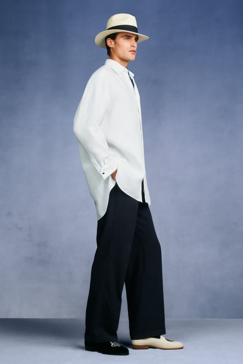 Model Parker Van Noord in a cream Panama hat with a navy band wears an oversized white linen tunic shirt over wide-leg navy trousers and embroidered velvet slippers
