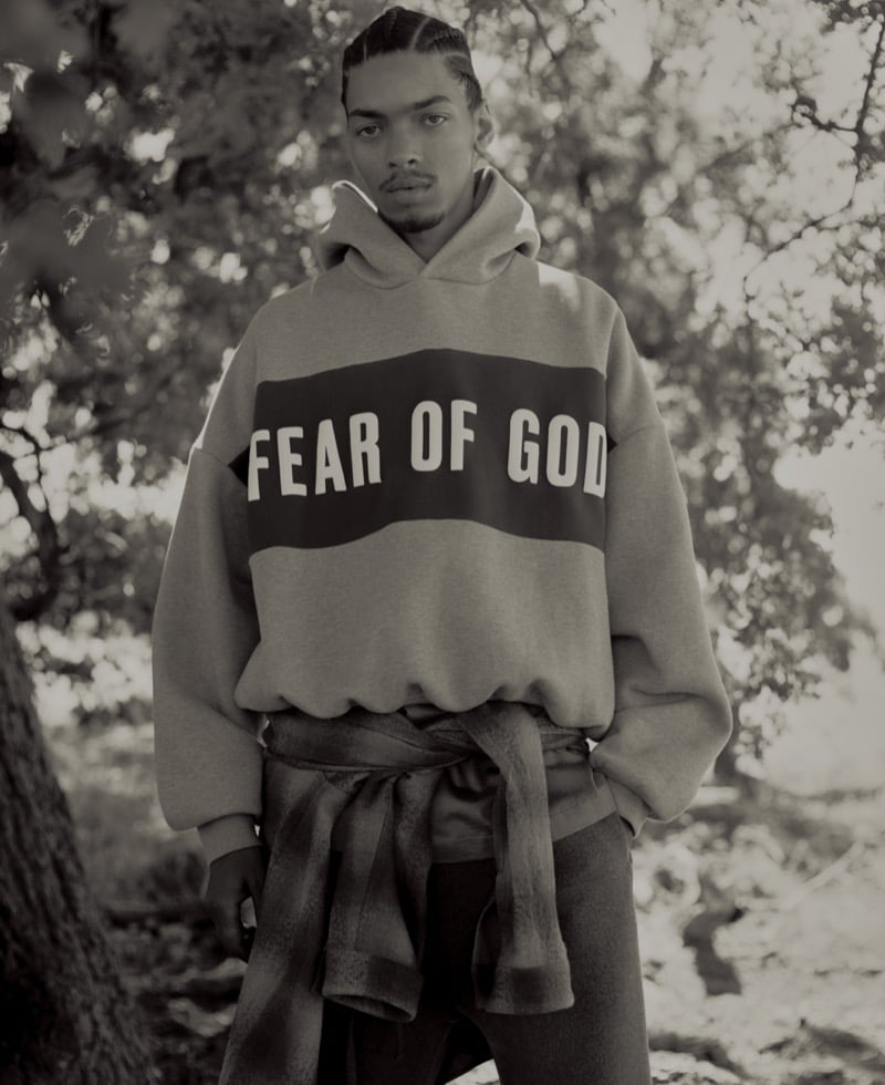 Black-and-white image of Omari Phipps in an oversized FEAR OF GOD logo hoodie with a flannel tied around the waist, standing among trees