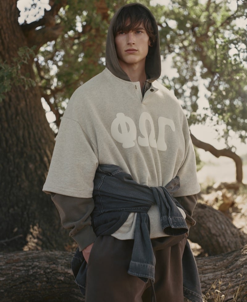 Sean Linden wears a heather short-sleeve sweatshirt with Greek FOG lettering over a full-zip hoodie, a denim trucker tied at the waist, and brown wide-leg pants