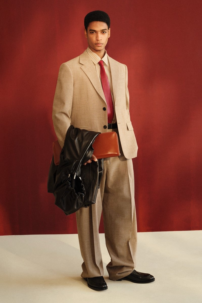 Male model in a tan wool single-breasted blazer over a brown striped shirt with a burgundy leather tie, paired with wide-leg taupe trousers and black leather loafers, holding a black leather coat and tan clutch against a red backdrop