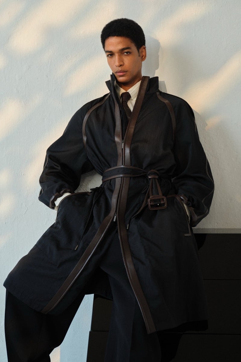 Male model in a black cotton trench coat with brown leather trim and a wraparound leather belt, layered over a cream shirt with a brown tie and dark trousers