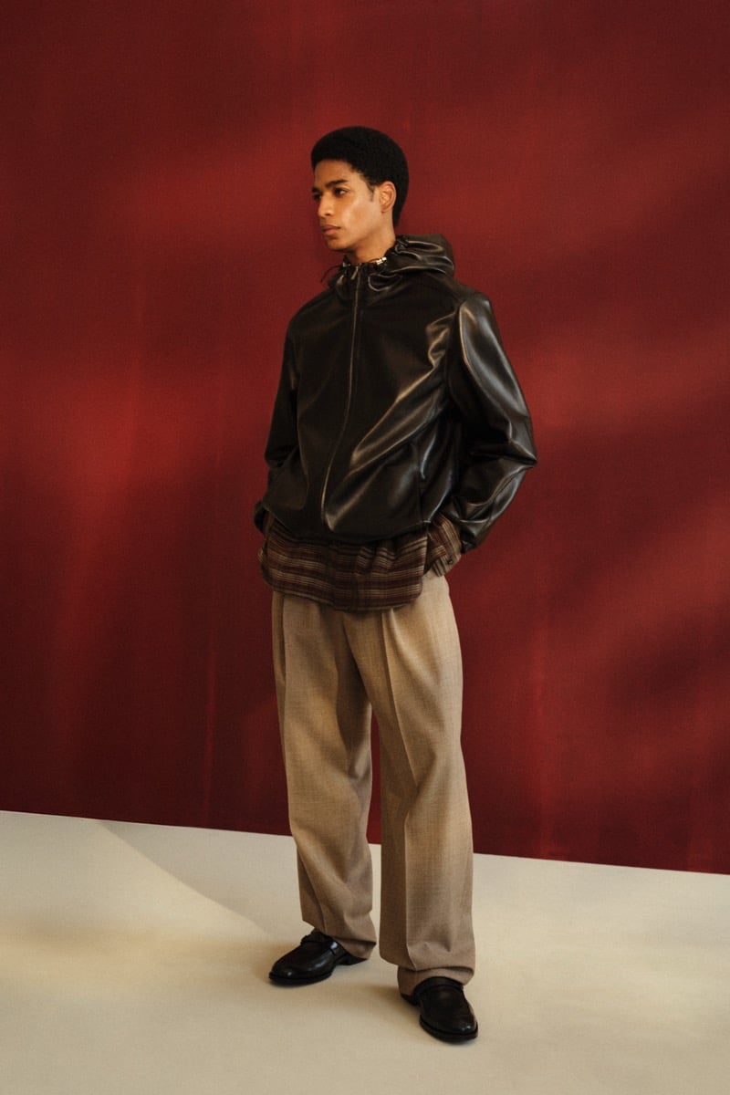 Male model in a black leather hooded zip bomber jacket over a brown plaid flannel shirt, paired with taupe wide-leg trousers and black leather loafers against a red backdrop