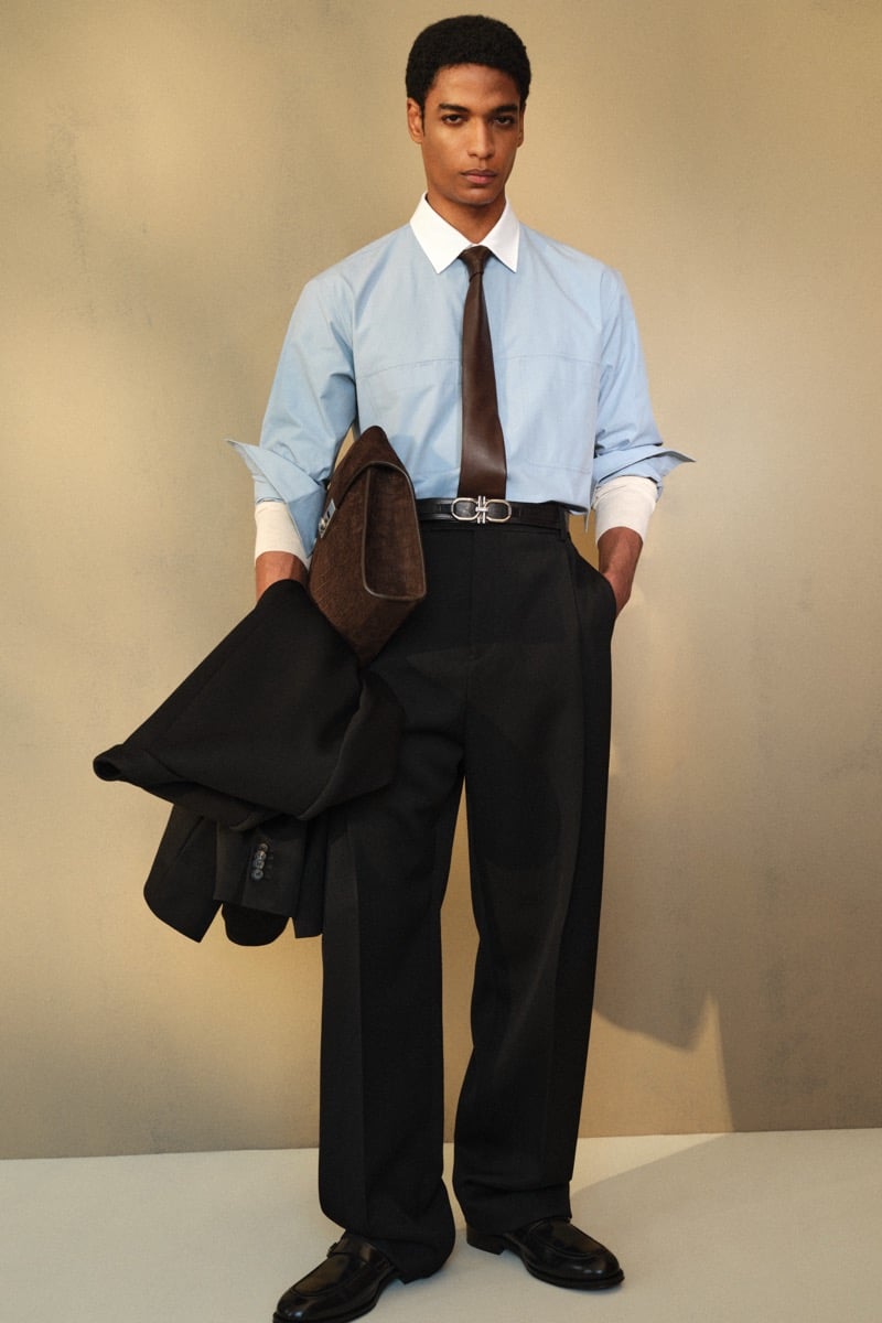 Male model in a light blue cotton dress shirt with a brown leather tie and silver Gancini buckle belt, paired with black wide-leg trousers and black leather loafers, holding a folded brown and black blazer
