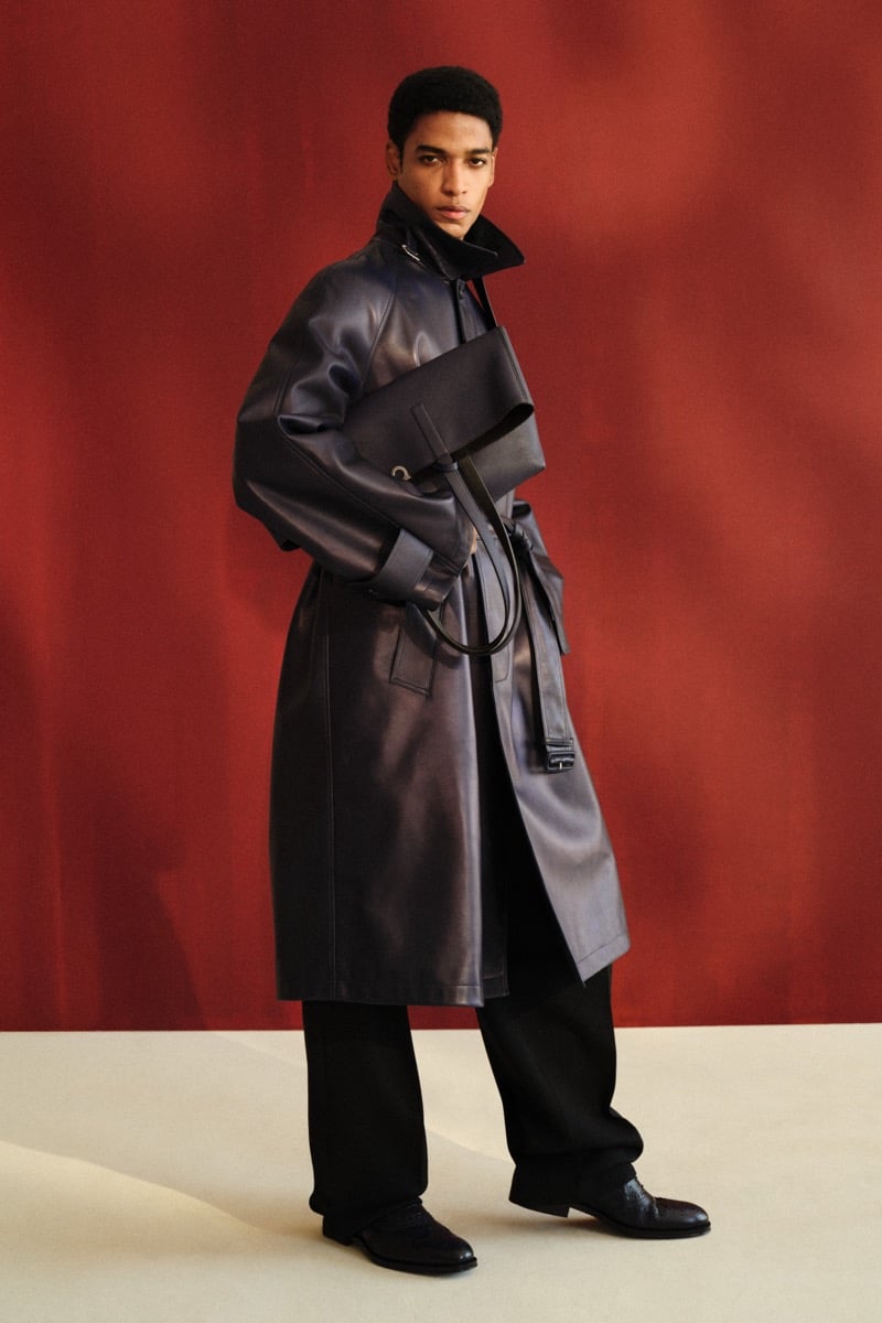 Male model in a navy leather trench coat with a stand collar and self-belt over black trousers and black loafers, carrying a structured black leather tote against a red backdrop