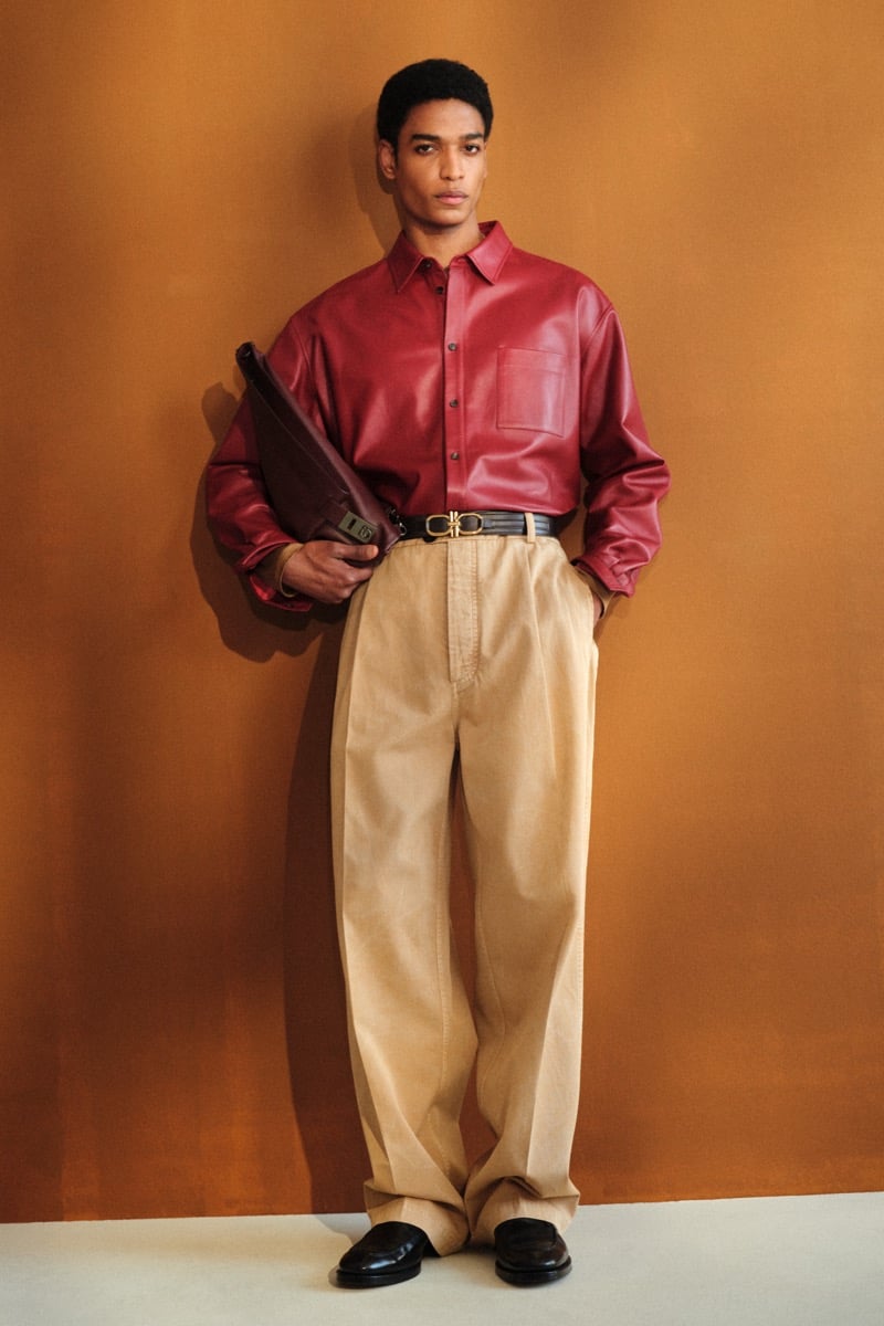 Male model in a burgundy leather button-down shirt tucked into high-waisted beige cotton trousers with a black leather Gancini buckle belt and black loafers, holding a burgundy leather clutch
