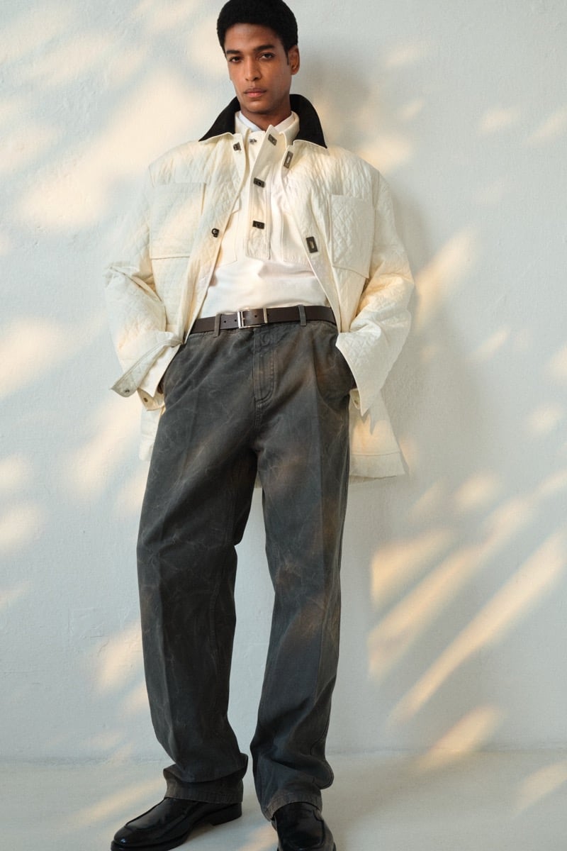 Male model in a cream quilted utility overshirt over a white mock-neck top with a brown leather belt, gray faded carpenter trousers, and black leather loafers
