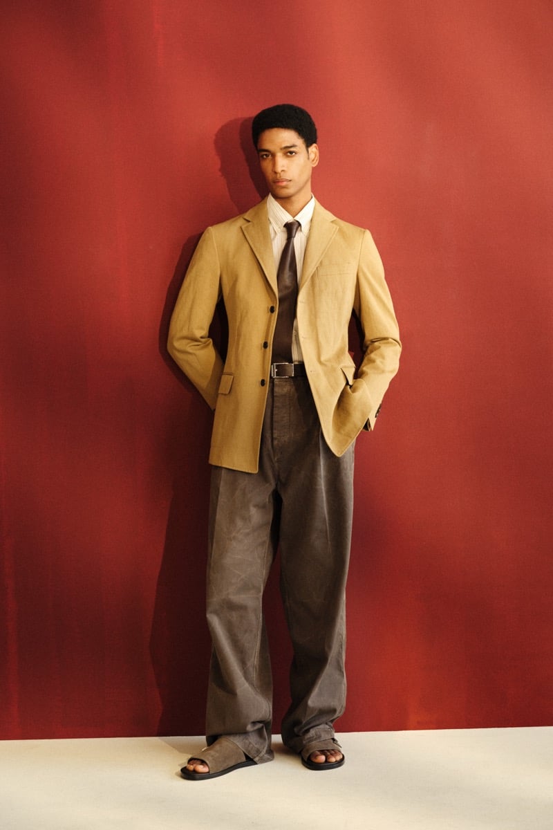 Male model in a tan suede single-breasted blazer over a brown striped shirt with a brown leather tie, paired with gray faded wide-leg trousers and dark suede sandals against a red backdrop