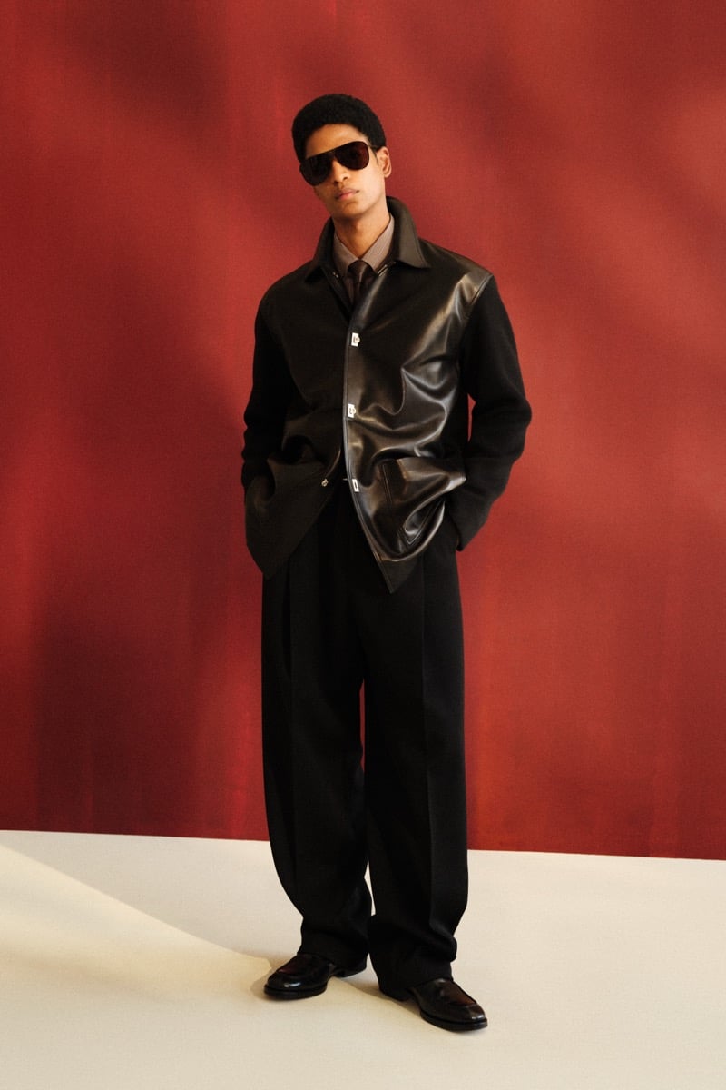 Male model in brown aviator sunglasses, a black wool jacket with a black leather panel front over a brown striped shirt and tie, paired with wide-leg black trousers and black leather loafers