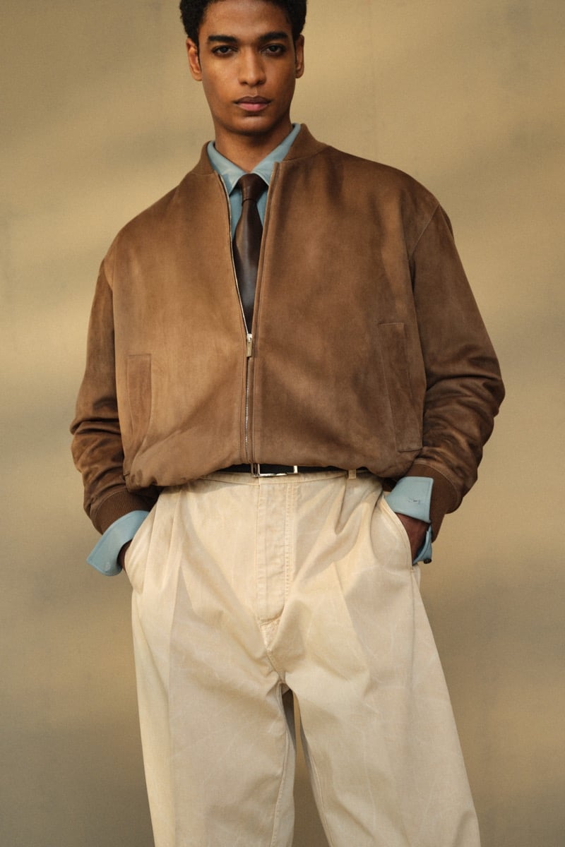 Male model in a tan suede bomber jacket over a light blue dress shirt with a brown leather tie, paired with cream pleated high-waisted trousers
