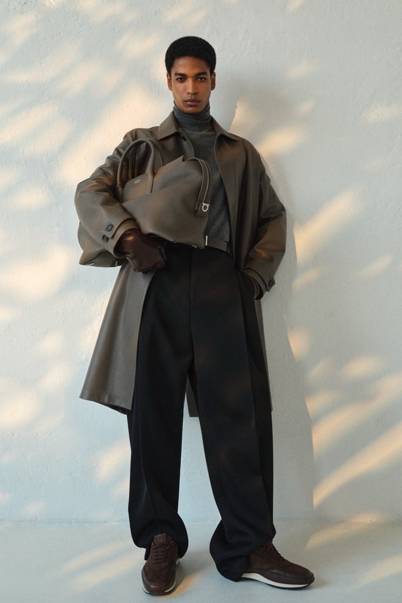 Male model in an olive leather trench coat over a gray turtleneck and black wide-leg trousers, with brown leather gloves and brown leather sneakers, holding a taupe leather tote with Gancini hardware