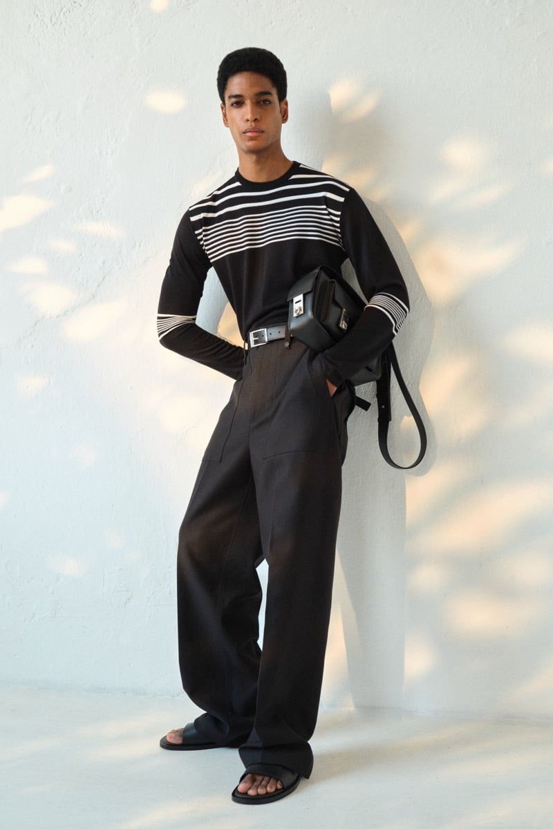 Male model in a black long-sleeve knit top with cream horizontal stripes across the chest, paired with wide-leg black trousers, a black leather belt with a crossbody bag, and black leather slide sandals