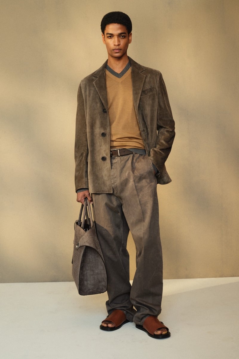 Male model in an olive suede single-breasted blazer over a camel V-neck sweater and gray T-shirt, paired with gray faded trousers and brown leather sandals, holding a gray croc-embossed leather tote