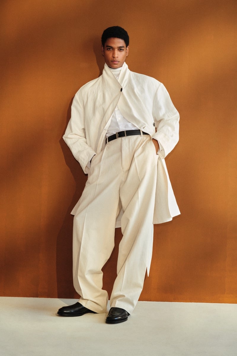 Male model in a cream linen wrap coat with stand collar over a white shirt and cream turtleneck, paired with cream pleated high-waisted trousers, a black belt, and black leather loafers