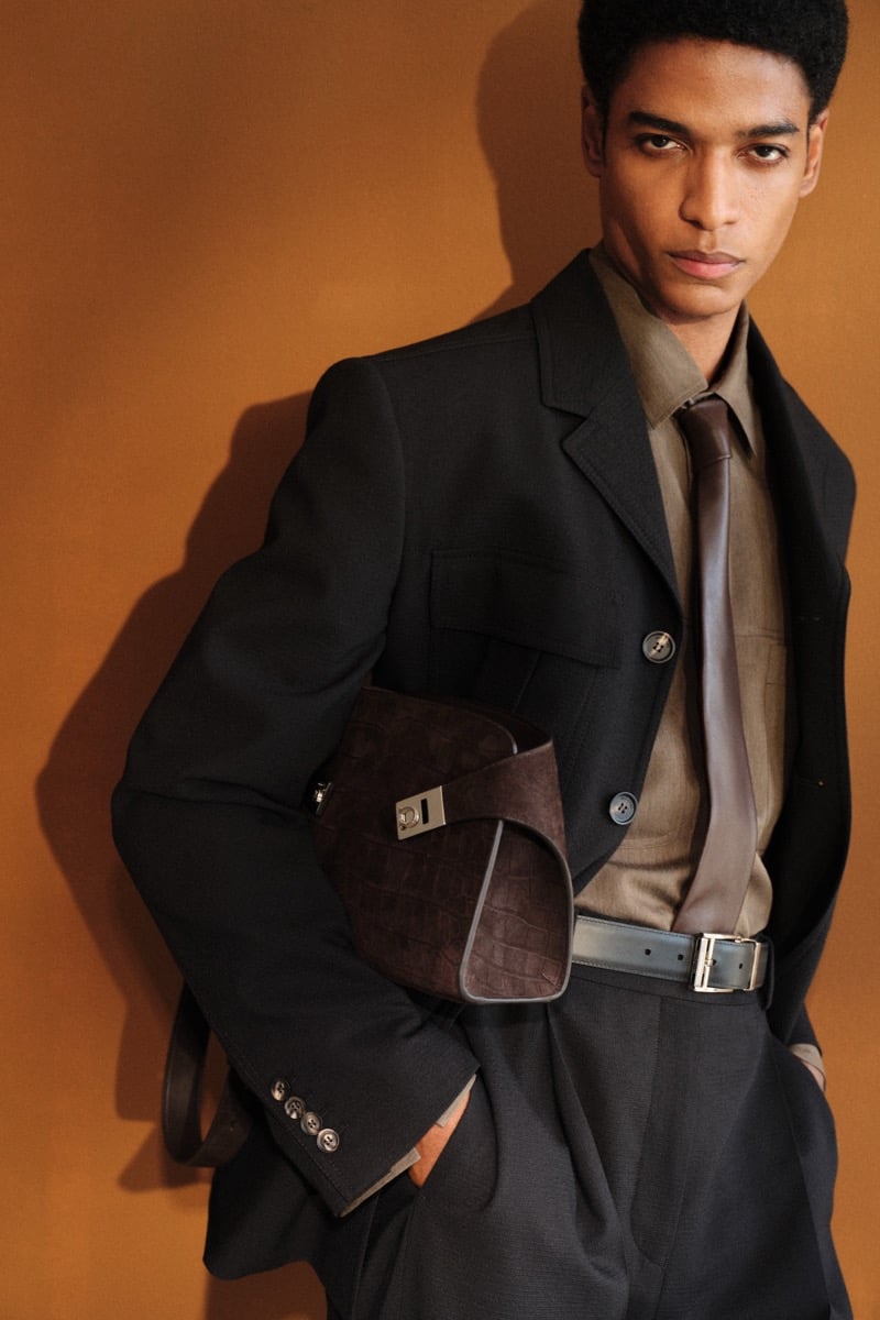Close-up of a male model in a black wool blazer with patch pockets over an olive silk shirt and brown silk tie, paired with a black leather belt and holding a brown croc-embossed leather clutch with a silver Gancini turn-lock