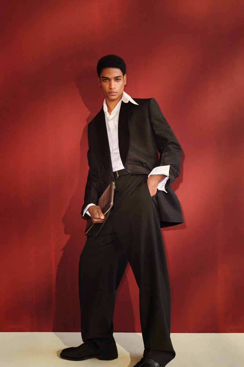 Male model in a cropped black tailored jacket with a quilted shawl lapel over a white shirt, paired with wide-leg black trousers and black leather loafers, holding a dark leather clutch against a red backdrop