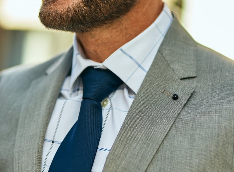Close view of a bearded man in a gray fresco wool suit jacket with a notch lapel and small lapel pin, paired with a windowpane-check shirt and a dark tie, the dry open weave visible at close range