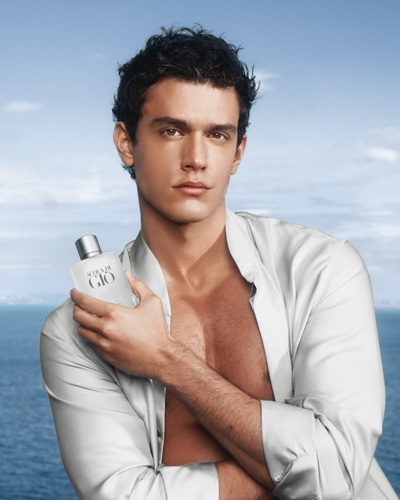 Xavier Serrano with tousled dark hair against a soft Mediterranean horizon, wearing an open pale silver silk shirt slipped off the shoulders and holding a frosted Giorgio Armani Acqua di Giò bottle against his bare chest with arms crossed