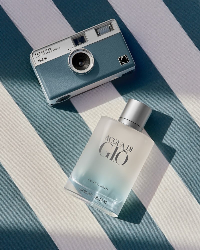 Overhead still life of the Giorgio Armani Acqua di Giò Eau de Toilette bottle beside a blue and white Kodak Ektar H35 half-frame film camera, arranged on a sunlit cabana towel striped in blue and ivory