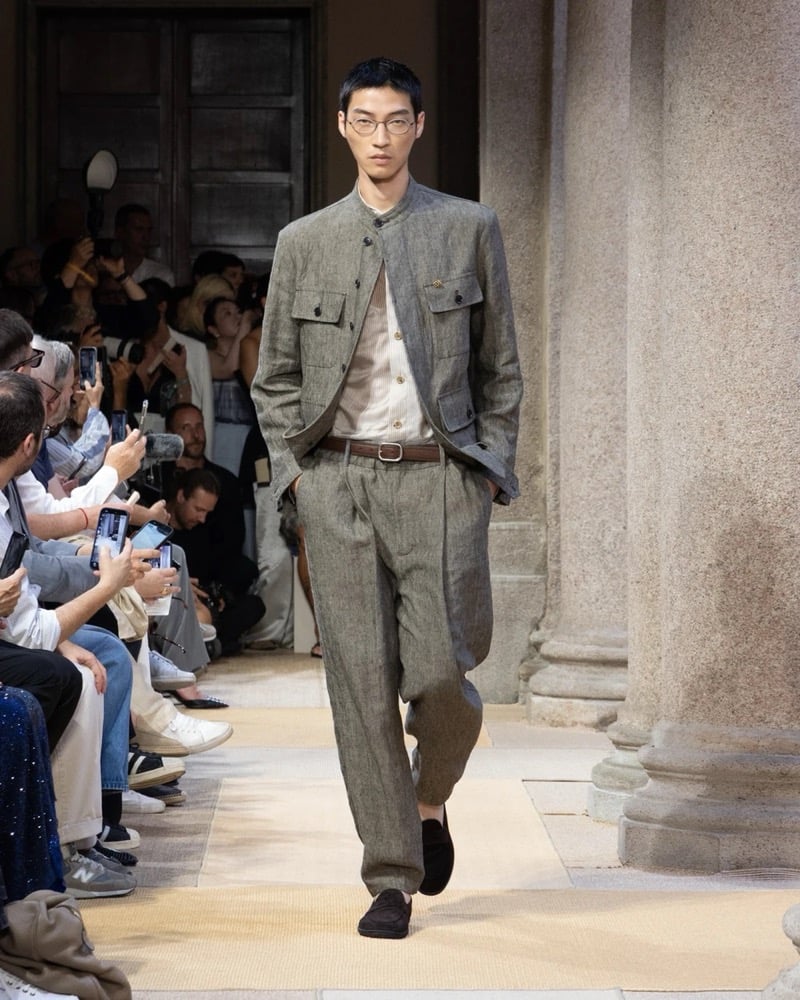 Model wears a band-collar linen jacket with chest patch pockets, layered over a soft shirt and worn with wide pleated linen trousers