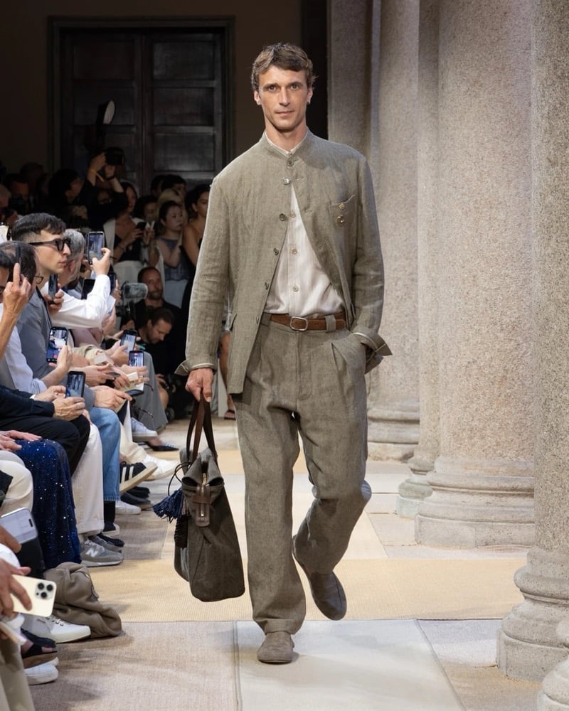 Model wears a relaxed band-collar linen jacket with a single chest pocket over a soft shirt, paired with wide pleated linen trousers