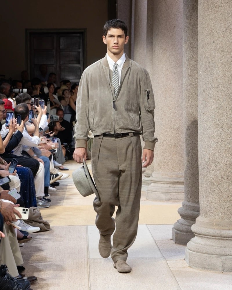 Model wears a soft suede bomber jacket with a zip front and a sleeve utility pocket, layered over a shirt and tie with wide pleated trousers