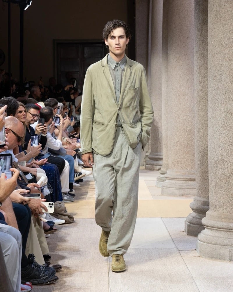 Model wears a soft suede single-button jacket over an open shirt, paired with wide pleated drawstring linen trousers