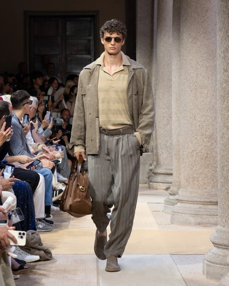 Model wears a relaxed linen safari jacket layered over a striped short-sleeve knit polo, paired with wide pleated pinstripe trousers