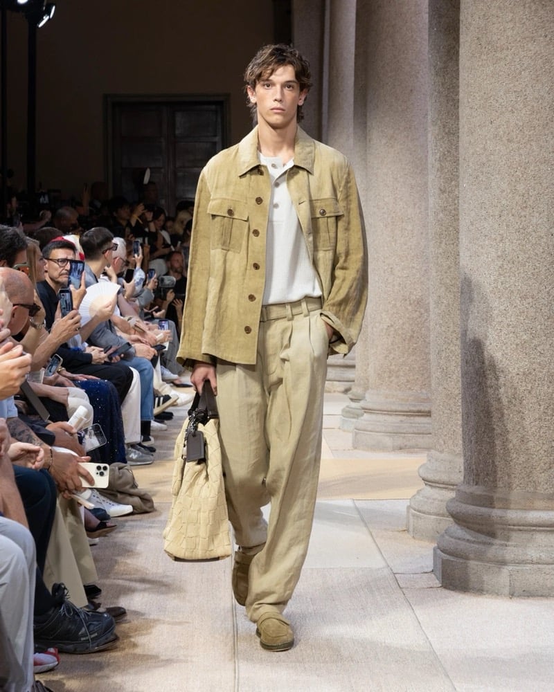 Model wears a relaxed suede shirt-jacket with twin chest pockets, layered over a soft polo and worn with wide pleated linen trousers