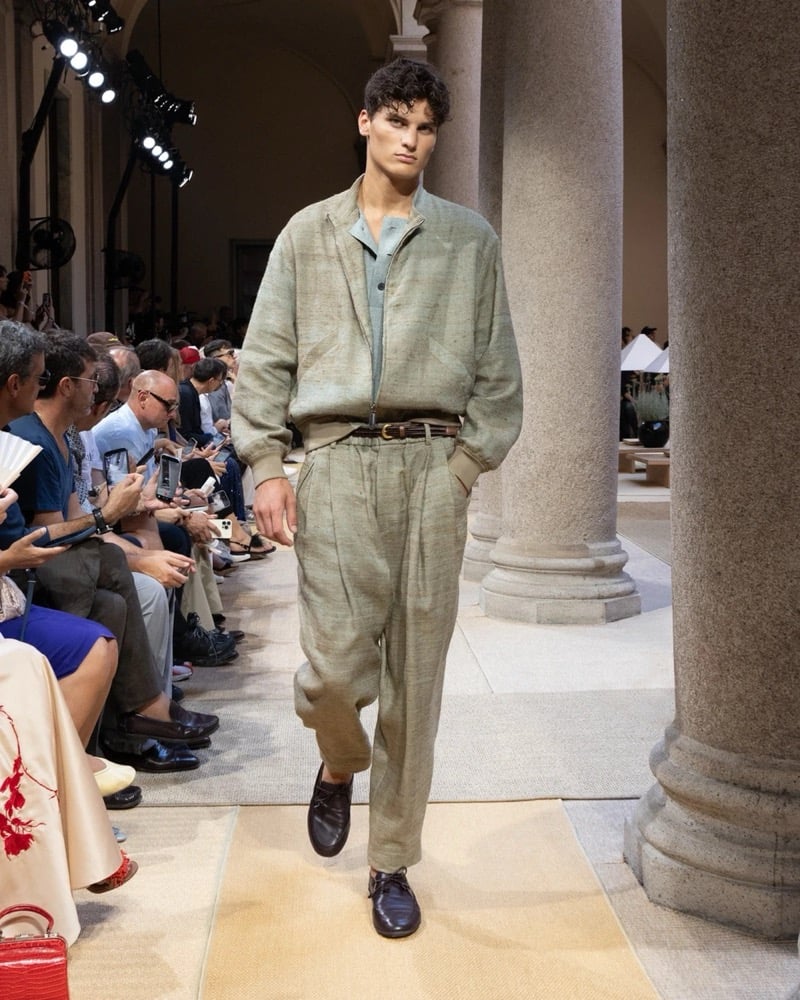 Model wears a soft linen blouson with banded cuffs and hem, layered over a half-buttoned shirt and worn with wide pleated trousers