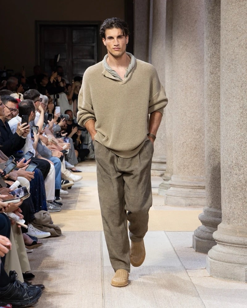 Model wears a chunky open-shawl-collar knit pullover layered over a soft scarf, paired with wide pleated linen trousers