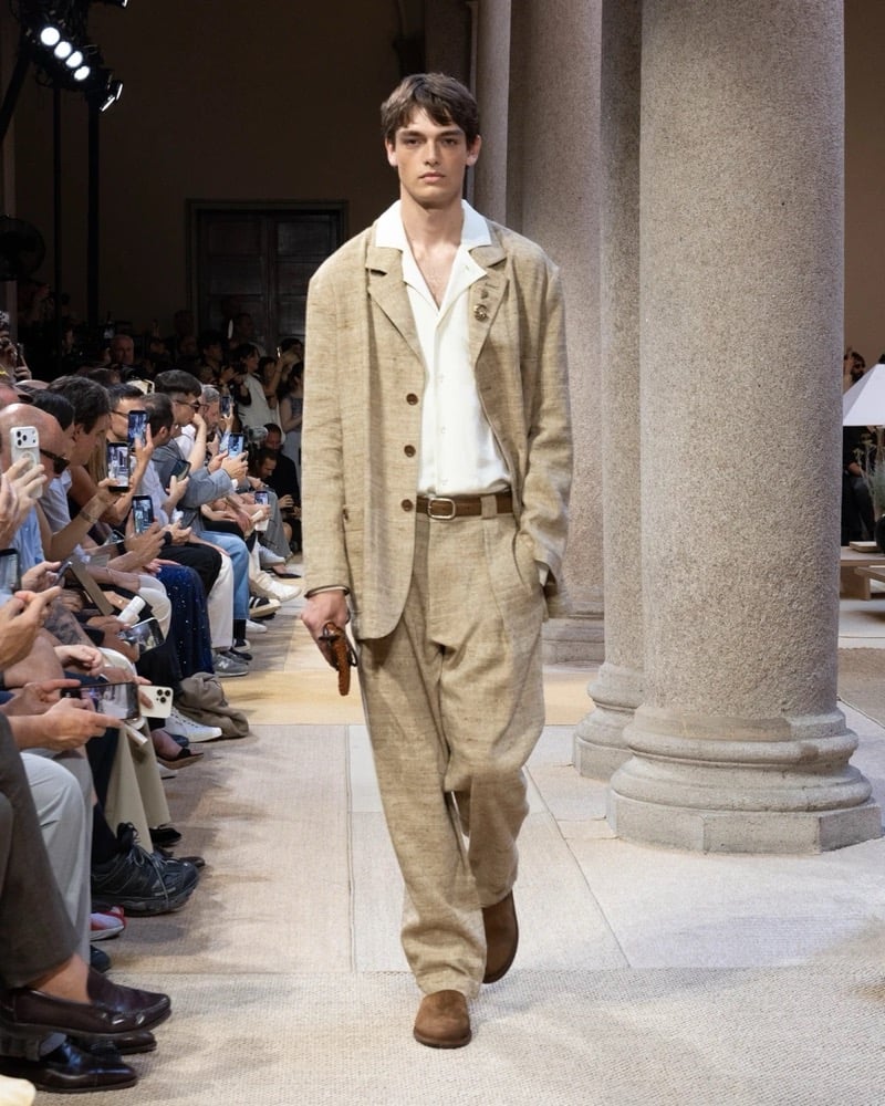 Model wears a four-button linen chore jacket with notch lapels and rounded pockets, layered over an open shirt with matching wide pleated trousers