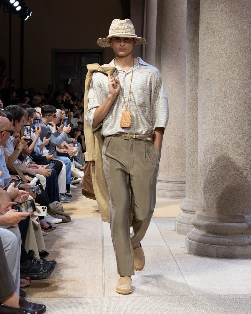 Model wears an oversized short-sleeve camp-collar striped knit polo paired with wide pleated silk trousers, a long draped coat over the arm