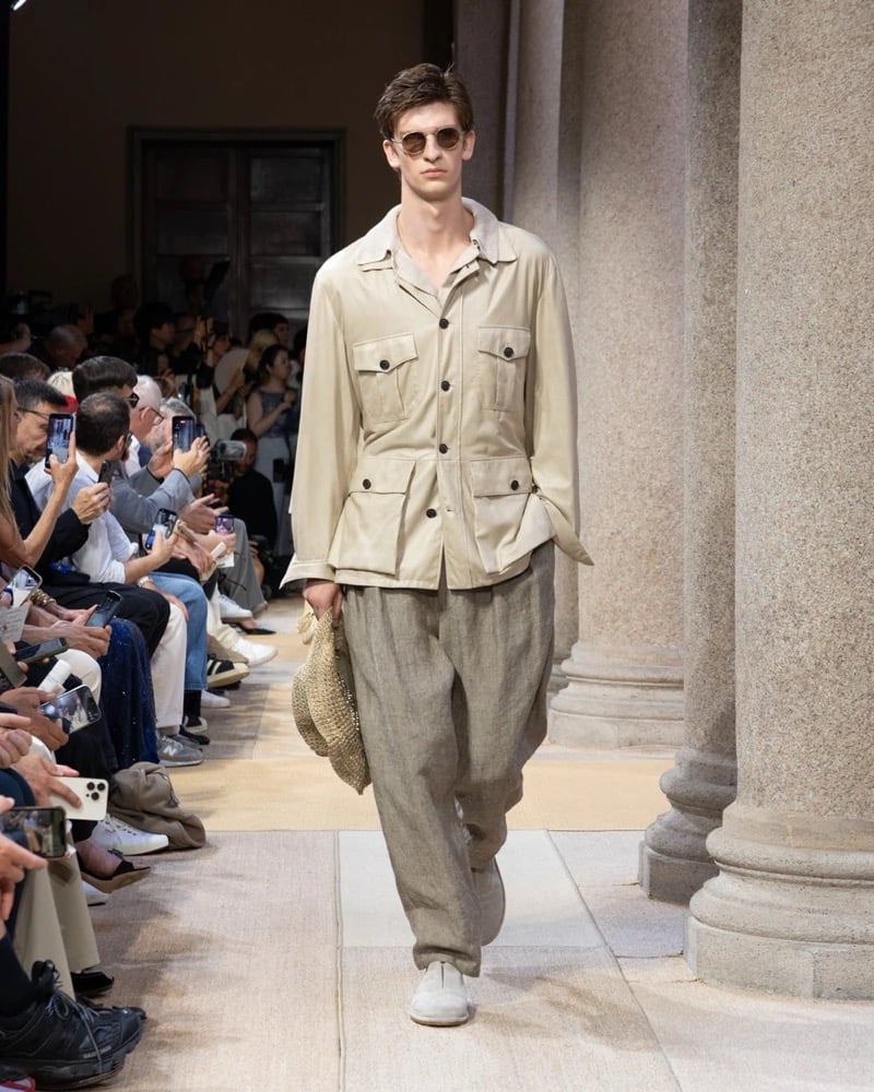 Model wears a relaxed safari-style linen jacket with four flap pockets, paired with wide pleated linen trousers