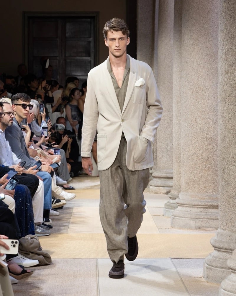 Model wears a relaxed single-button linen blazer with patch pockets layered over a soft open shirt, paired with wide pleated linen trousers