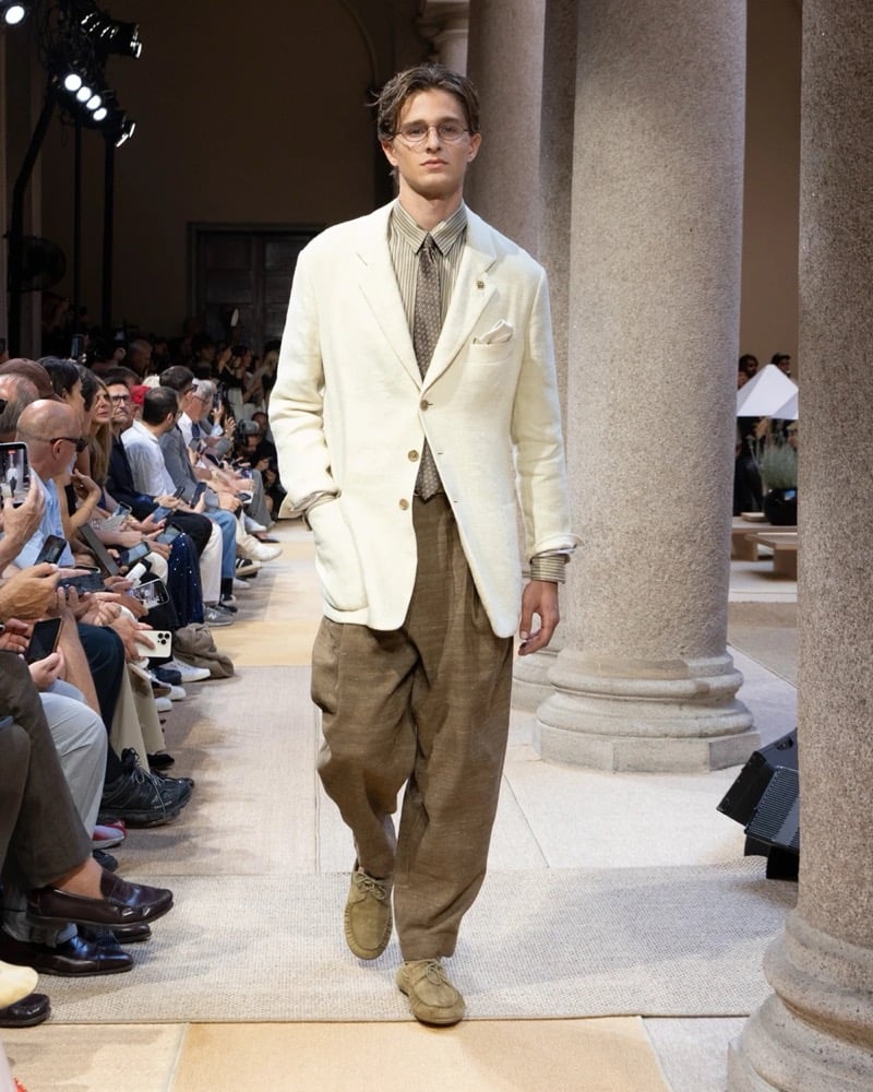 Model wears a relaxed single-breasted linen blazer over a striped shirt with a printed tie and waistcoat, paired with wide pleated linen trousers