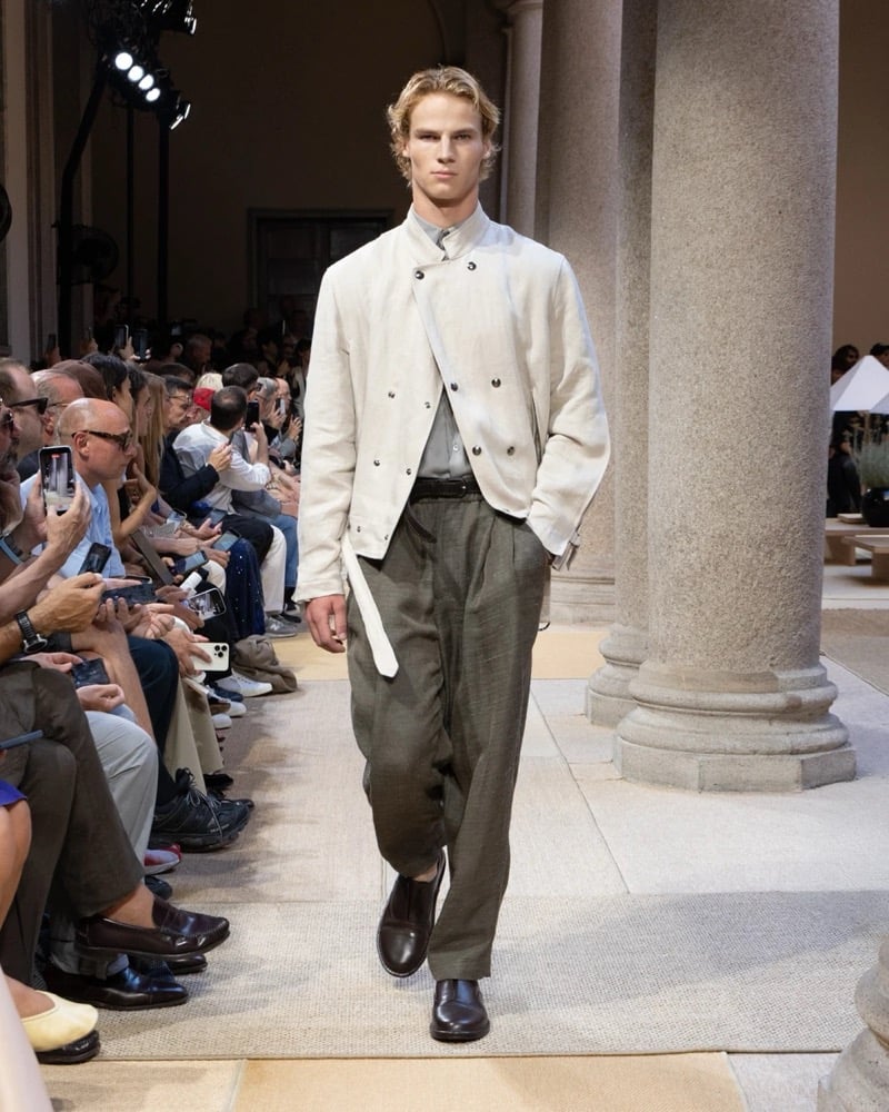Model wears a cropped band-collar linen jacket with a double-button placket and tied front, layered over a shirt with wide pleated trousers