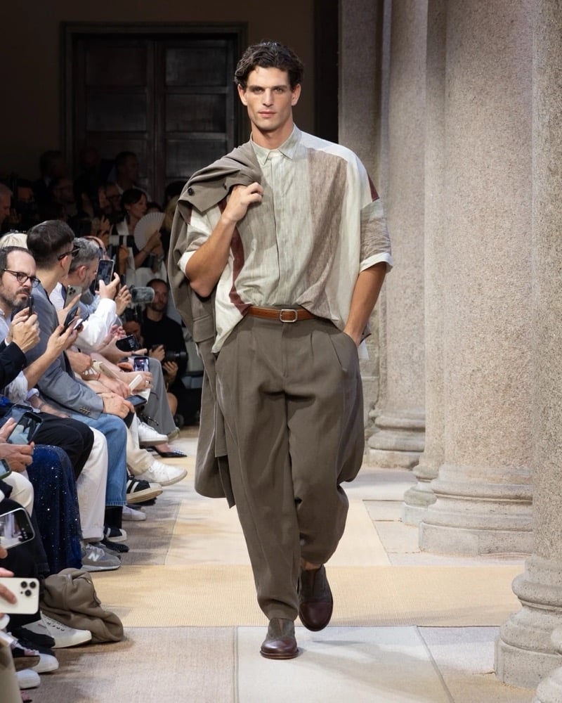 Model wears a draped short-sleeve patchwork-paneled shirt tucked into wide pleated linen trousers, an outer jacket carried over the shoulder