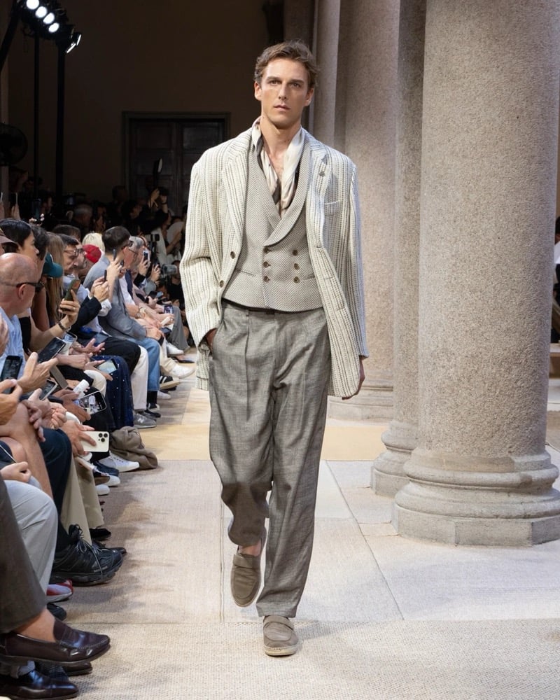 Model wears a pinstripe linen blazer with notch lapels layered over a double-breasted shawl-collar waistcoat and open shirt, paired with wide pleated trousers