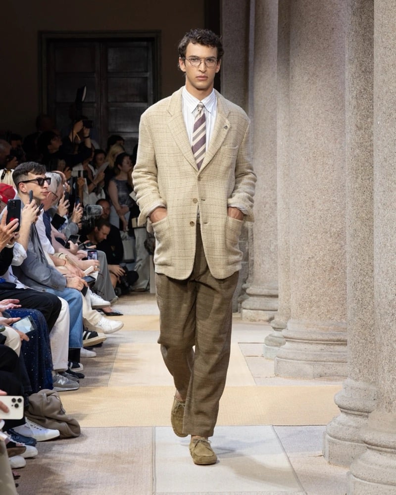 Model wears a textured boucle linen blazer with notch lapels over a shirt and striped tie, paired with wide pleated linen trousers