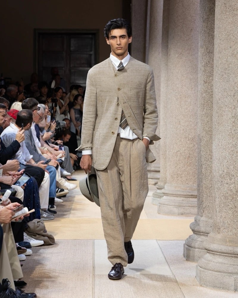 Model wears an asymmetric-buttoned band-collar linen jacket over a shirt and striped tie, paired with wide pleated linen trousers