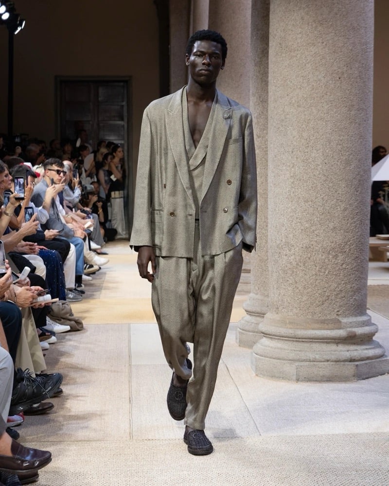 Model wears an oversized double-breasted linen blazer with peak lapels and patch pockets, layered over a shawl-collar waistcoat with wide pleated trousers